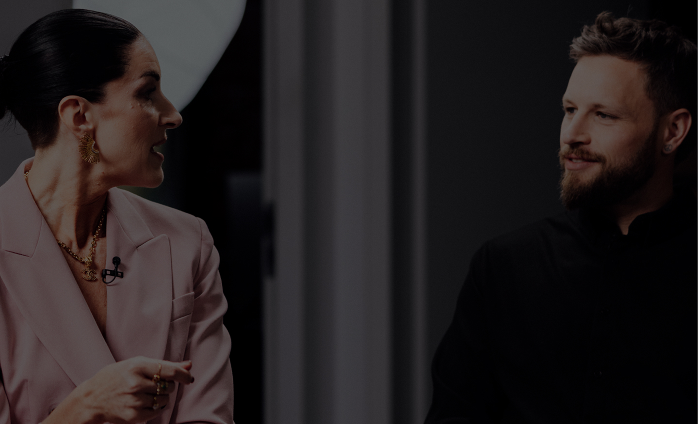 Zwei Personen im Gespräch in einem Studio: links eine Frau im rosa Blazer, rechts ein Mann in Schwarz